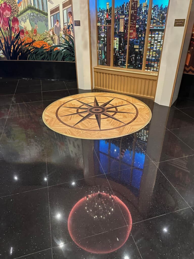 A decorative compass rose is set into a shiny black floor, reflecting lights and a red circular glow. Behind it, murals depict flowers, people, and a city skyline through tall windows.