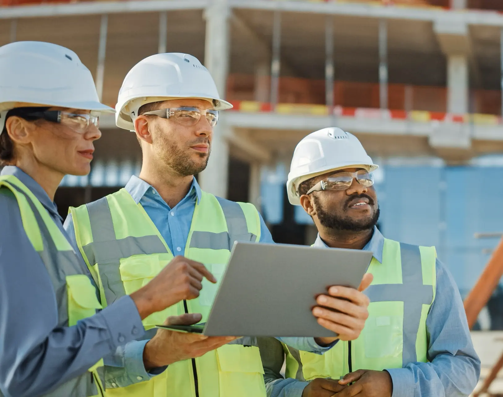 Three construction workers wearing safety vests, hard hats, and safety glasses stand at a building site, looking at a laptop together and discussing the project. A partially built structure is visible in the background.
