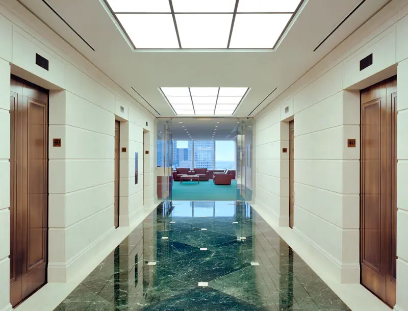 A modern building hallway with marble floors, two elevators on each side, and a seating area with large windows and red chairs at the far end. Bright ceiling lights illuminate the space.