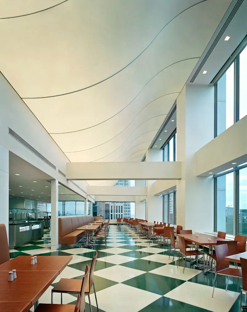 Spacious, modern cafeteria with brown tables and chairs, green and white checkered floor, large windows, and a wavy, cream-colored ceiling. Natural light fills the room, creating an open and inviting atmosphere.
