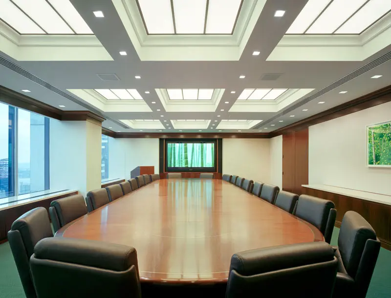 A large, empty boardroom with a long oval wooden table surrounded by black leather chairs, bright ceiling lights, large windows, and a screen displaying green scenery at the far end.