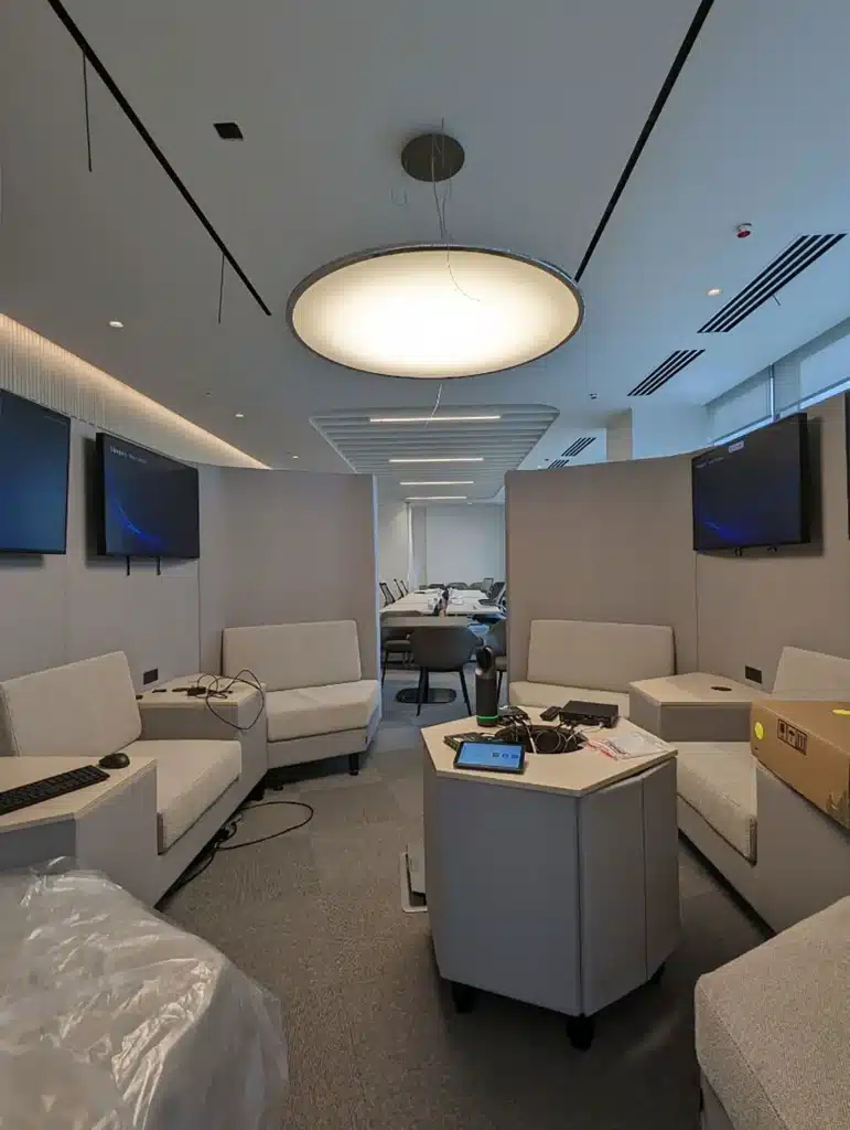 A modern office space with gray sofas arranged in a square, monitors mounted on the walls, a central table with cables and devices, and a large circular ceiling light. The room is clean and minimalistic.