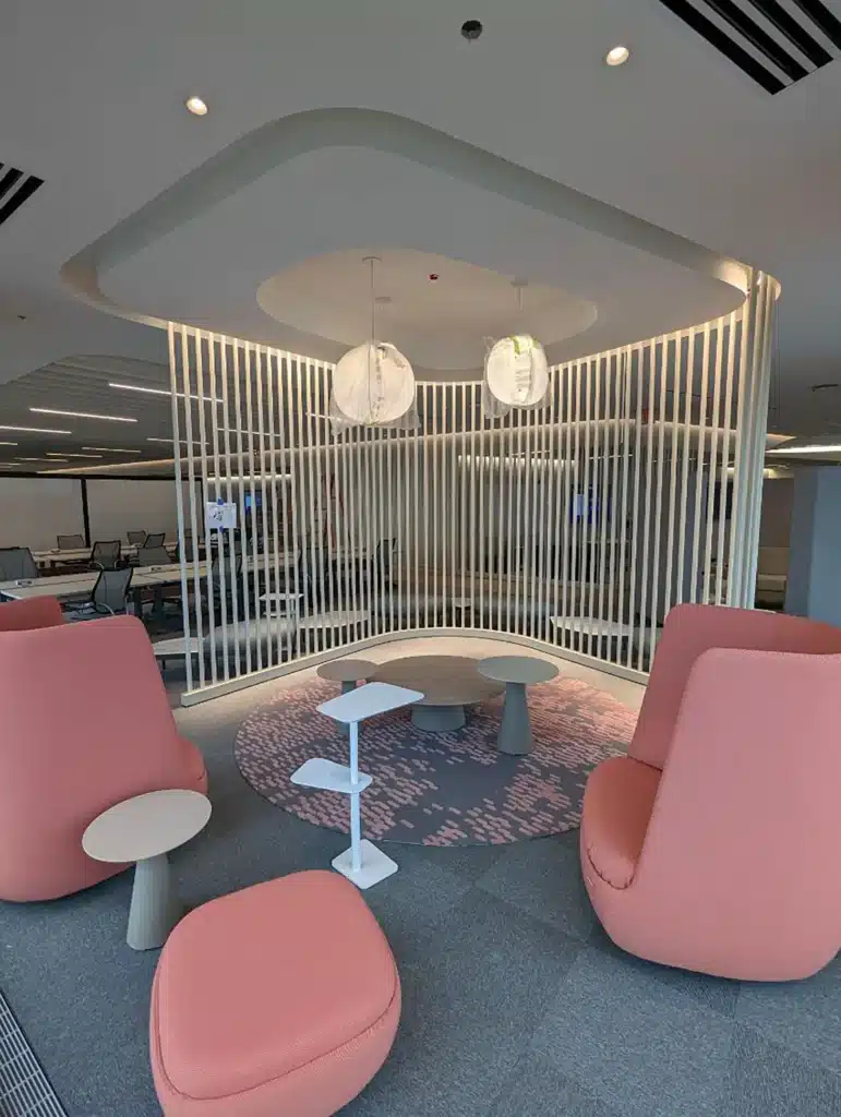 Modern office lounge area with pink cushioned chairs, white side tables, a round rug, two pendant lights, and a decorative white slat partition. Open workspace with desks and chairs visible in the background.