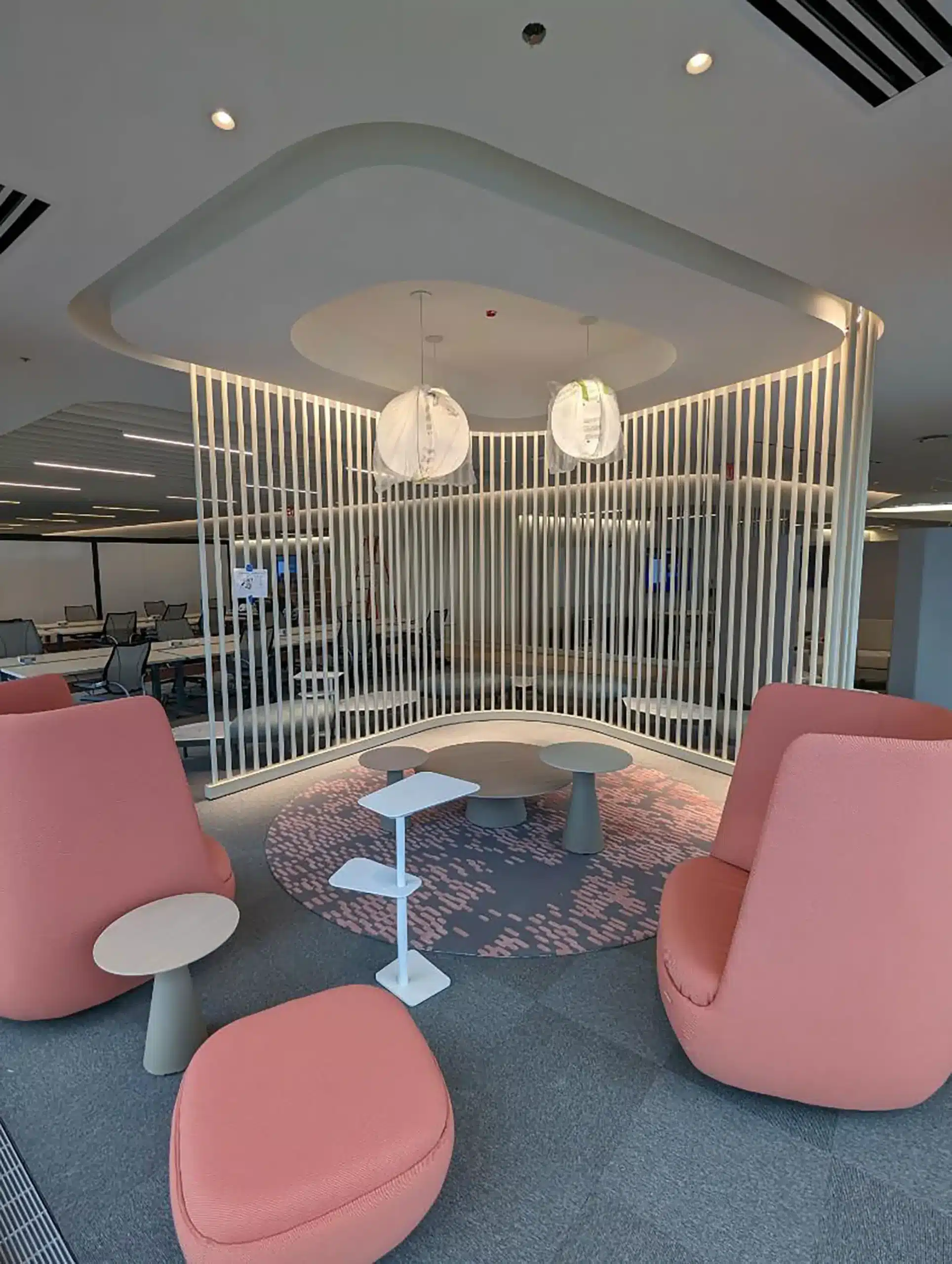 Modern office lounge area with pink cushioned chairs, white side tables, a round rug, two pendant lights, and a decorative white slat partition. Open workspace with desks and chairs visible in the background.