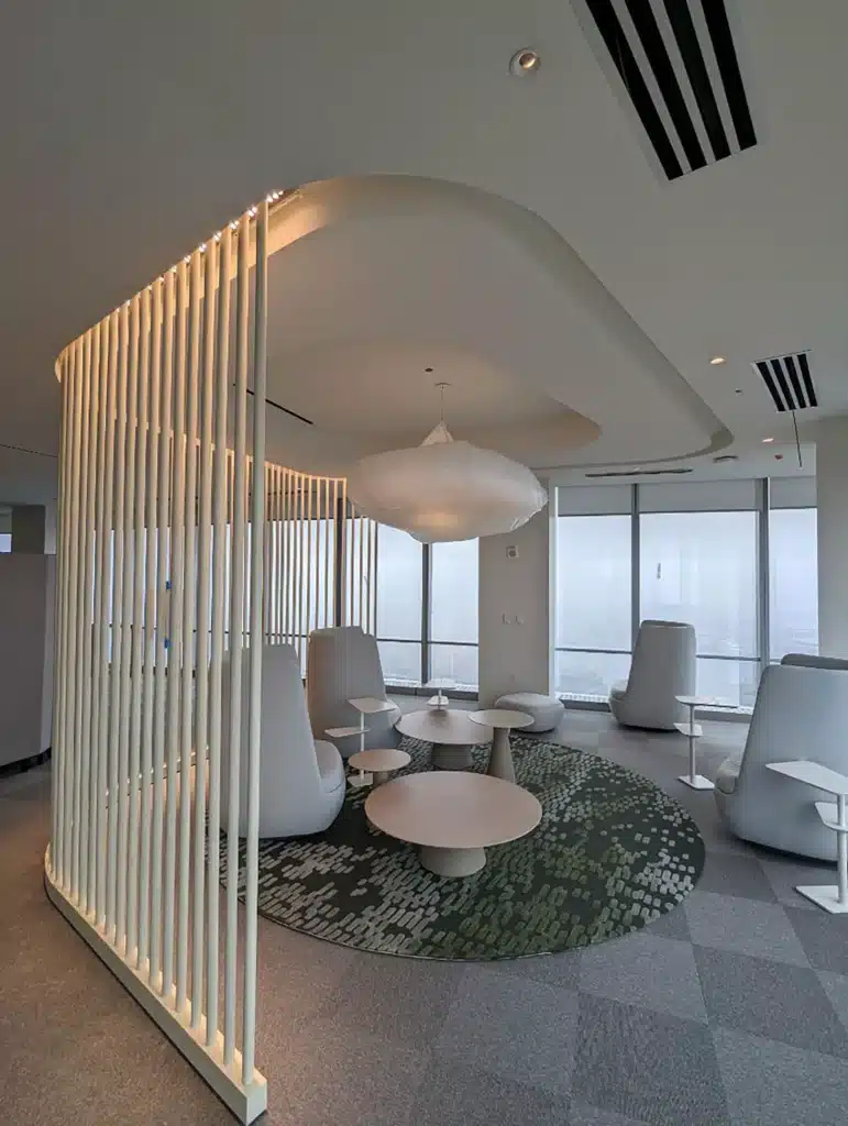 A modern lounge with white chairs, round tables, a green patterned rug, and a cloud-like ceiling light, separated by vertical slats, with large windows providing natural light.