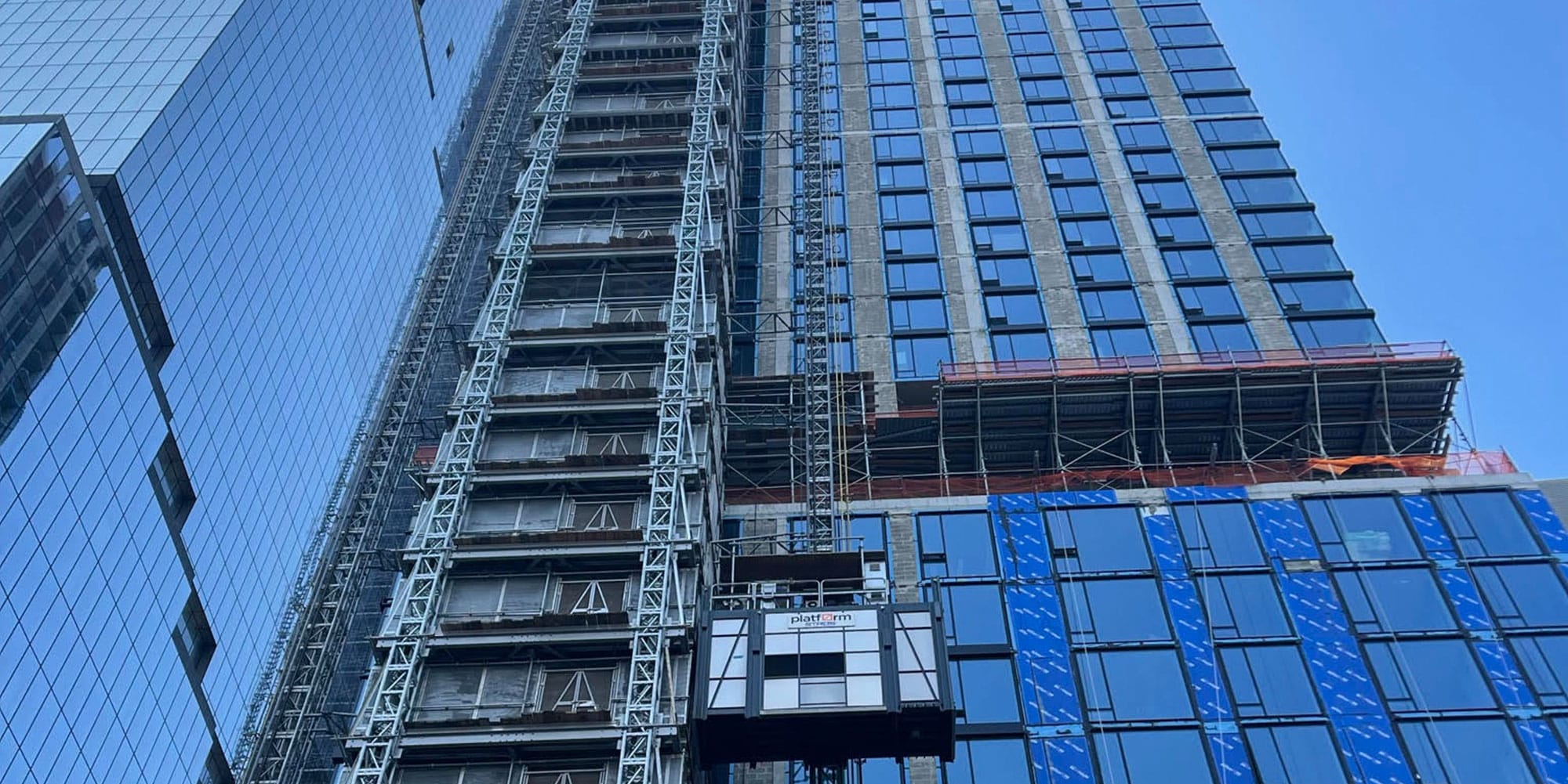 A construction elevator is attached to the side of a tall glass skyscraper under construction, with scaffolding and blue sky reflected in the building’s windows.