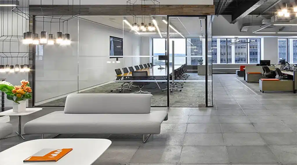 Modern office interior with gray sofas, a white coffee table holding an orange notebook, glass-walled conference room with chairs, and large windows overlooking city buildings. Industrial lighting and exposed ceiling beams are visible.