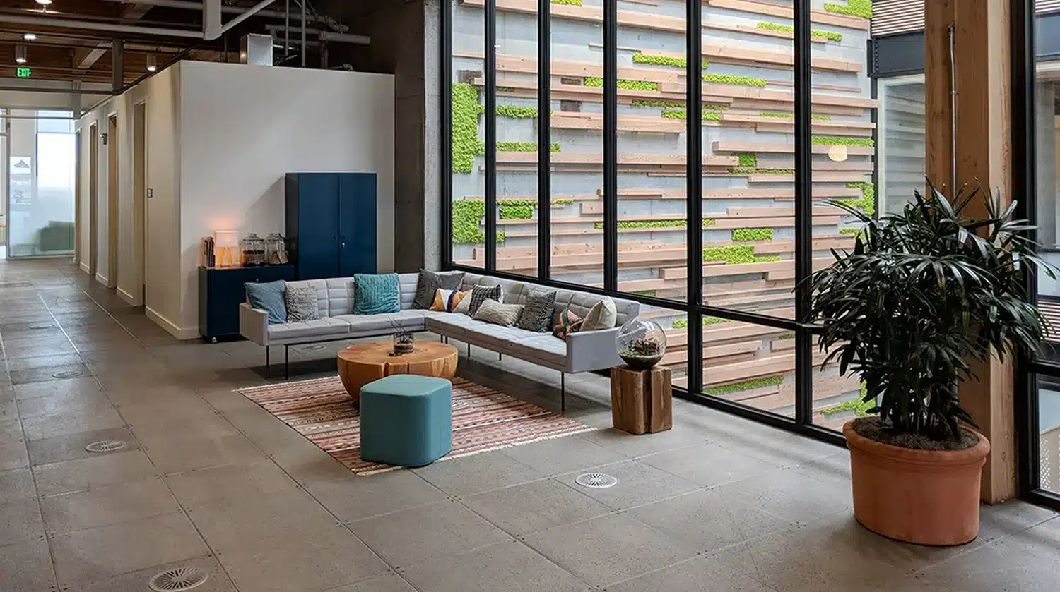 Modern office lounge with a gray sectional sofa, colorful pillows, small tables, a striped rug, potted plant, and large windows overlooking a building wall decorated with wood panels and greenery.