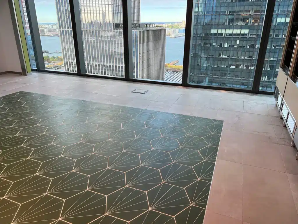 A modern, high-rise office with large windows, a view of a river and city buildings, and a floor featuring large beige tiles with a section of dark green geometric patterned tiles.