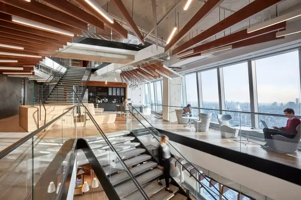 Modern office interior with large windows, glass railings, and wooden ceiling accents. People are seated and working in lounge chairs, while one person walks down the staircase. Cityscape visible through the windows.