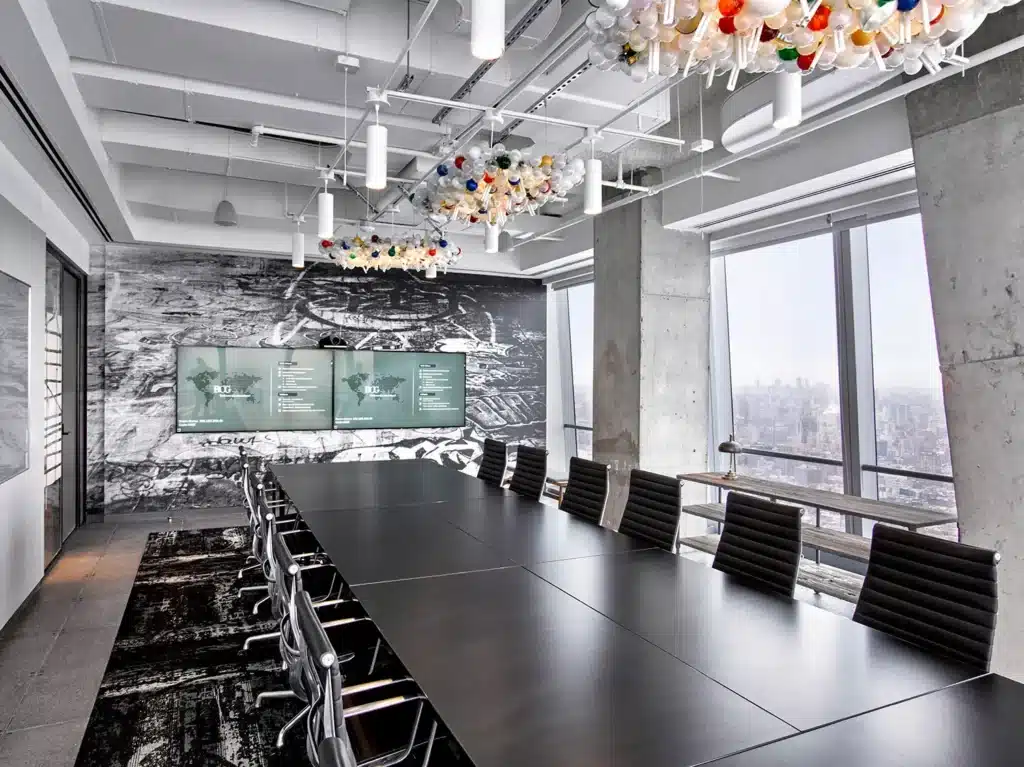 A modern conference room with a long black table, black chairs, large windows with a city view, three wall-mounted screens, a monochrome mural, concrete columns, and colorful ceiling light fixtures.