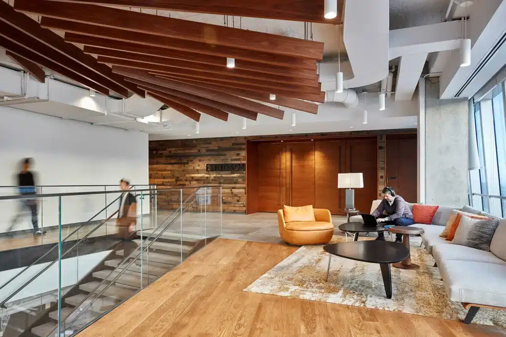 Modern office space with wood panel accents, staircase, glass railing, and seating area featuring a sofa, an orange chair, a coffee table, and a person working on a laptop. Another person descends the stairs.