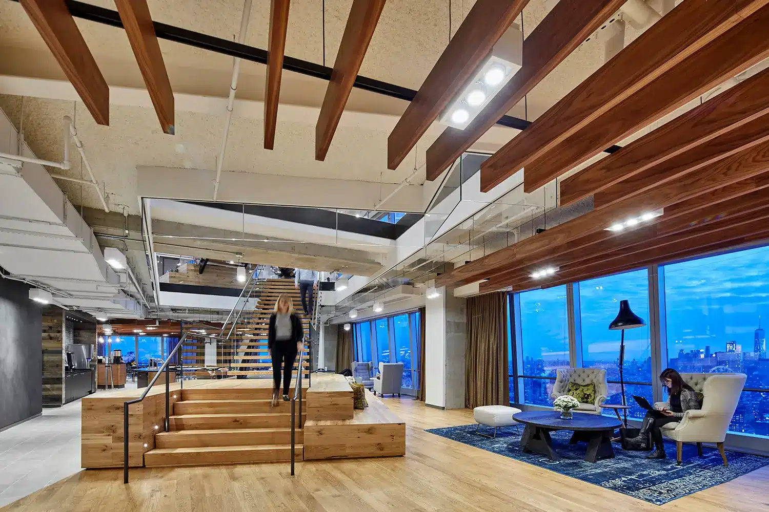Modern office space with wooden floors and beams, open seating areas, large windows overlooking a cityscape at dusk, and two people—one walking down stairs and another seated on a couch using a laptop.