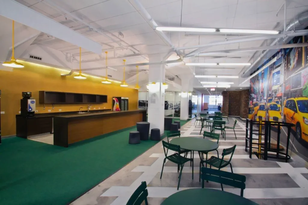 A modern, open office space with green chairs and tables, a kitchen area with a long counter, yellow accent wall, hanging lights, and a large mural of yellow taxis along one wall. The floor has green and white sections.