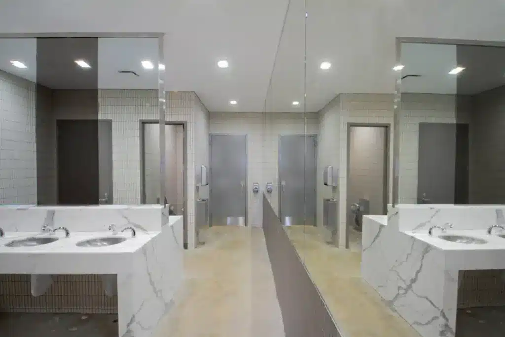 A modern public restroom with light tile walls, large mirrors, two stainless steel sinks in a marble countertop, and gray stall doors. The mirrors create a symmetrical reflection of the space.