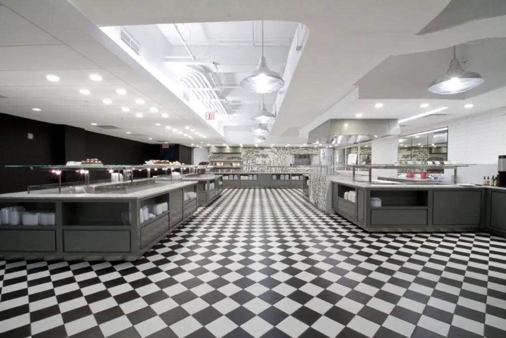 A spacious, modern commercial kitchen with black and white checkered floor tiles, stainless steel counters, open shelves, and bright overhead lighting. The area is empty and clean, ready for use.