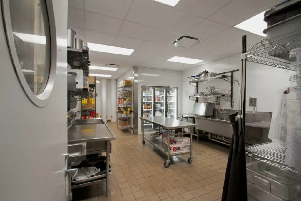 A spacious commercial kitchen with stainless steel counters, shelving units lined with supplies, a large refrigerator, and tiled floors under bright overhead lighting.