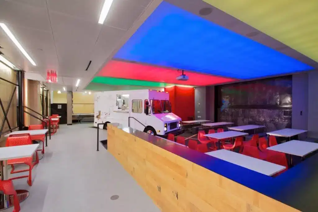 Modern cafeteria with red chairs and white tables, featuring a food truck indoors. The ceiling is lit with blue, yellow, and red lights, creating a vibrant atmosphere. A wooden divider separates seating areas.