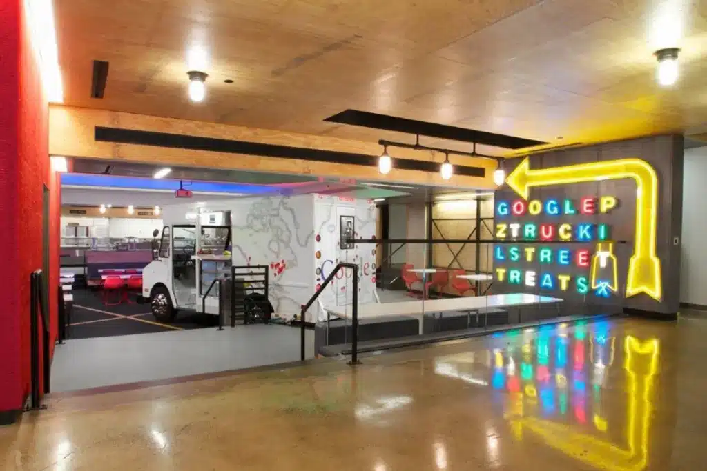 A modern office space with a white food truck indoors, tables and chairs nearby, and a large neon sign with colorful letters spelling “GOOGLE ZTRUCK STREET TREATS” and an arrow pointing to the truck.