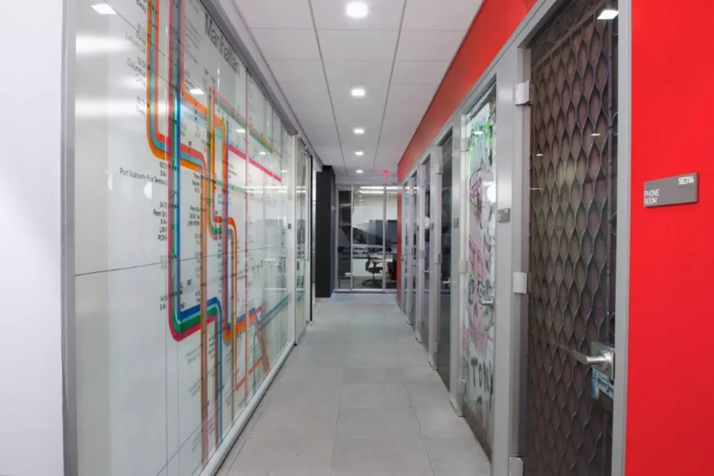 A modern office hallway with white walls, glass doors, and a large colorful map or diagram on the left wall; the ceiling has recessed lights and the right wall features a bright red accent.