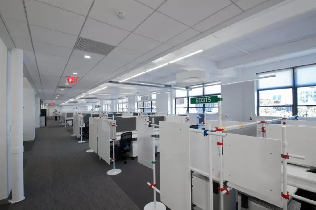 A modern open-plan office with rows of white cubicles, swivel chairs, computers, and partition panels. Large windows line one wall, letting in natural light. The ceiling has recessed lighting and ventilation panels.