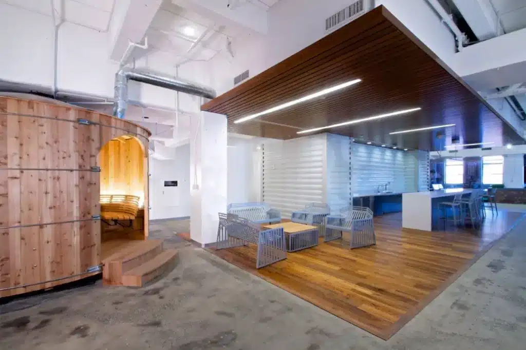 Modern office lounge with wood and metal furniture, a wooden pod-like meeting room on the left, and an open kitchen with bar seating in the back. Exposed ducts and natural light create an industrial yet inviting atmosphere.