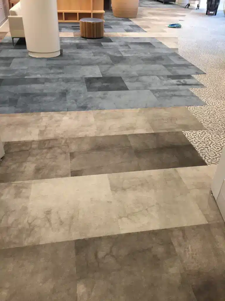 A floor with different sections of patterned carpet tiles in shades of gray, blue, and beige. In the background, there is a round ottoman and some furniture. A piece of blue cloth is on the floor in the distance.
