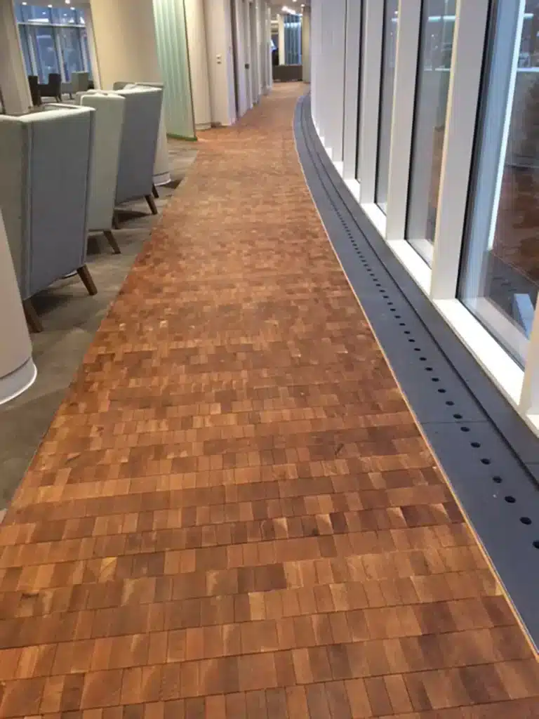 A corridor with large windows on one side and high-backed chairs on the other, featuring a floor made of small, square wooden tiles arranged in a grid pattern.