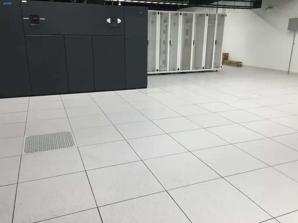 A clean, spacious server room with white tiled floors, a large dark-colored server unit on the left, and several empty white server racks against the back wall. Some cardboard boxes are stacked in the corner.