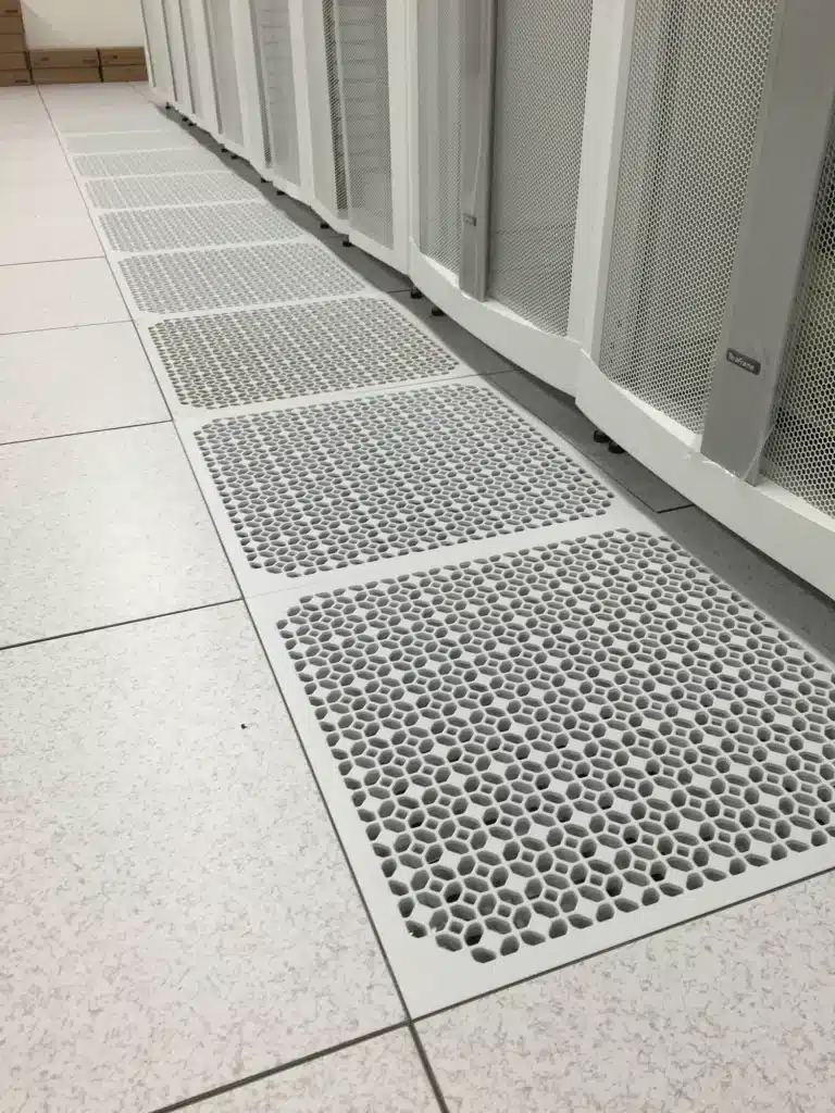 A row of perforated floor panels with hexagonal holes is installed next to white mesh cabinets in a clean, modern data center or server room environment.