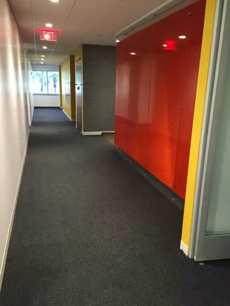 A hallway in an office building with dark carpet, white and yellow walls, and a section of bright orange glossy panels. There are ceiling lights and an exit sign visible at the far end near a glass door.