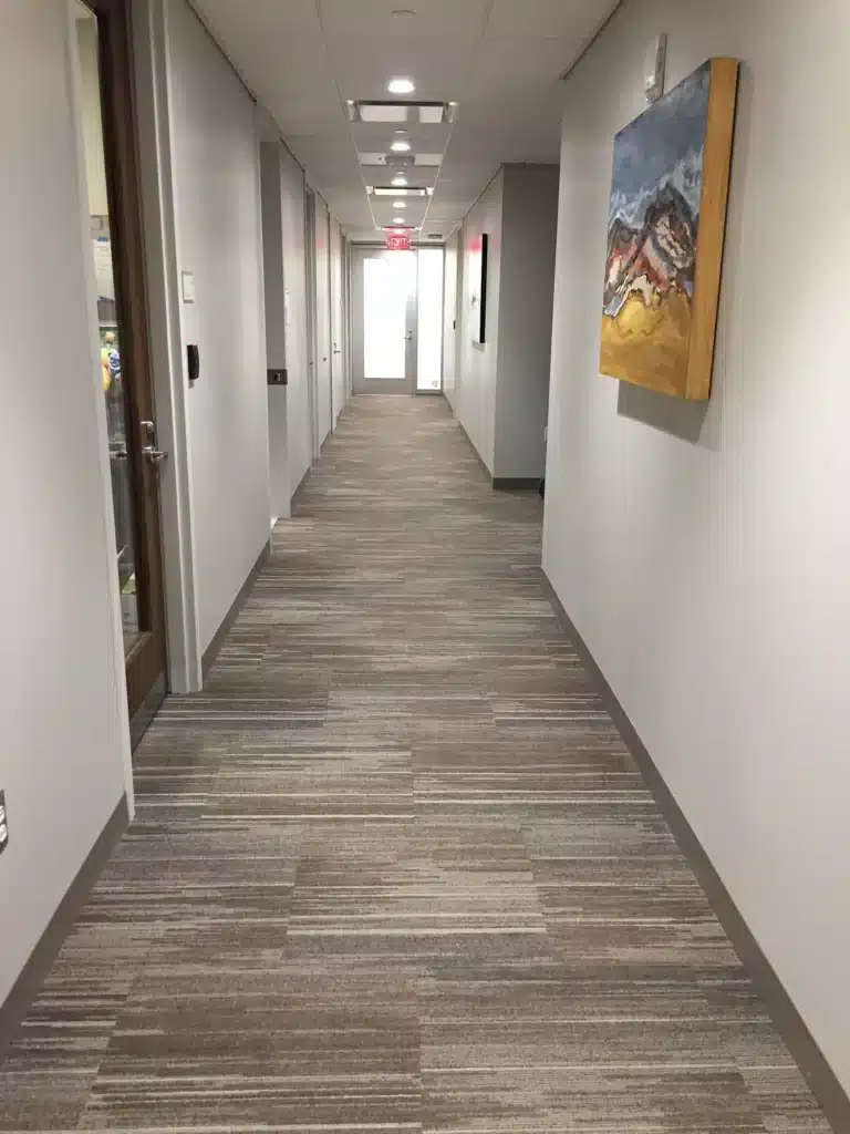 A long, well-lit hallway with beige carpet and white walls, featuring a colorful abstract painting on the right wall and office doors on the left. A bright exit is visible at the far end of the corridor.