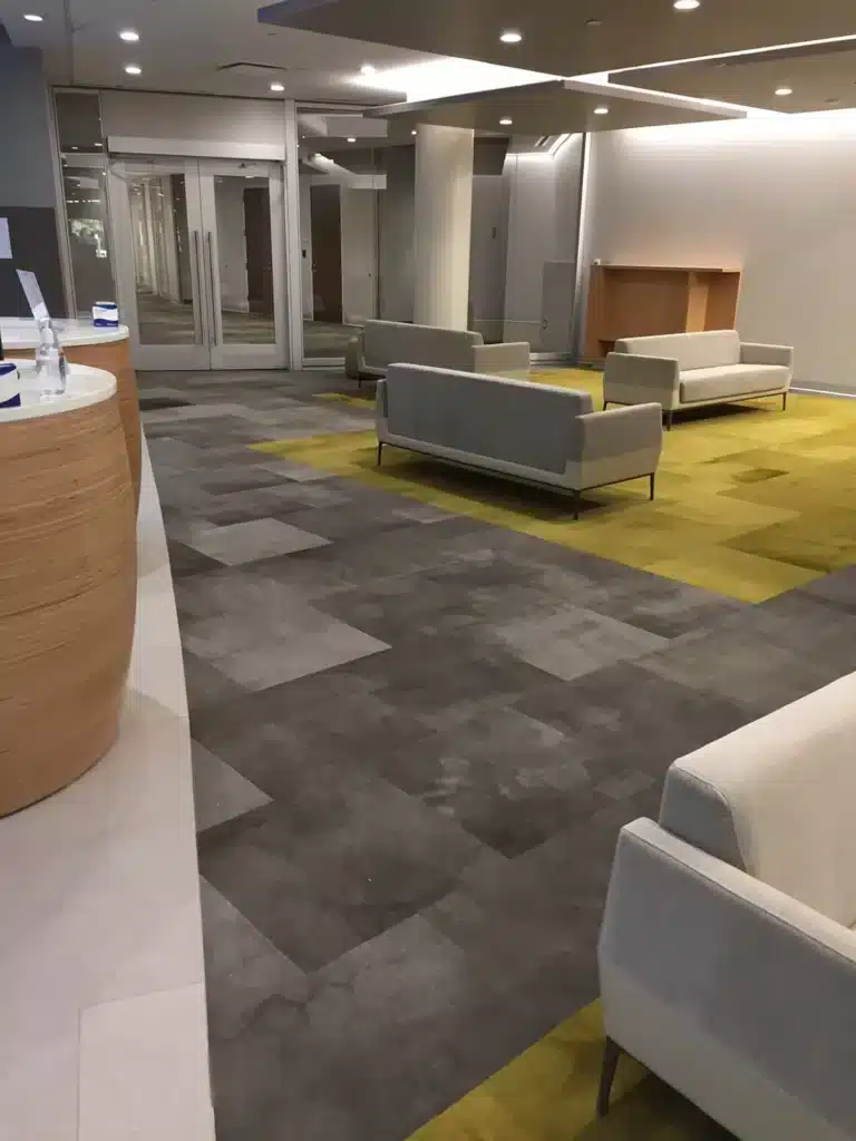 Modern office lobby with gray and yellow patterned carpeting, several light gray couches, wood-paneled reception desk on the left, and glass doors in the background. The area is brightly lit with overhead lights.