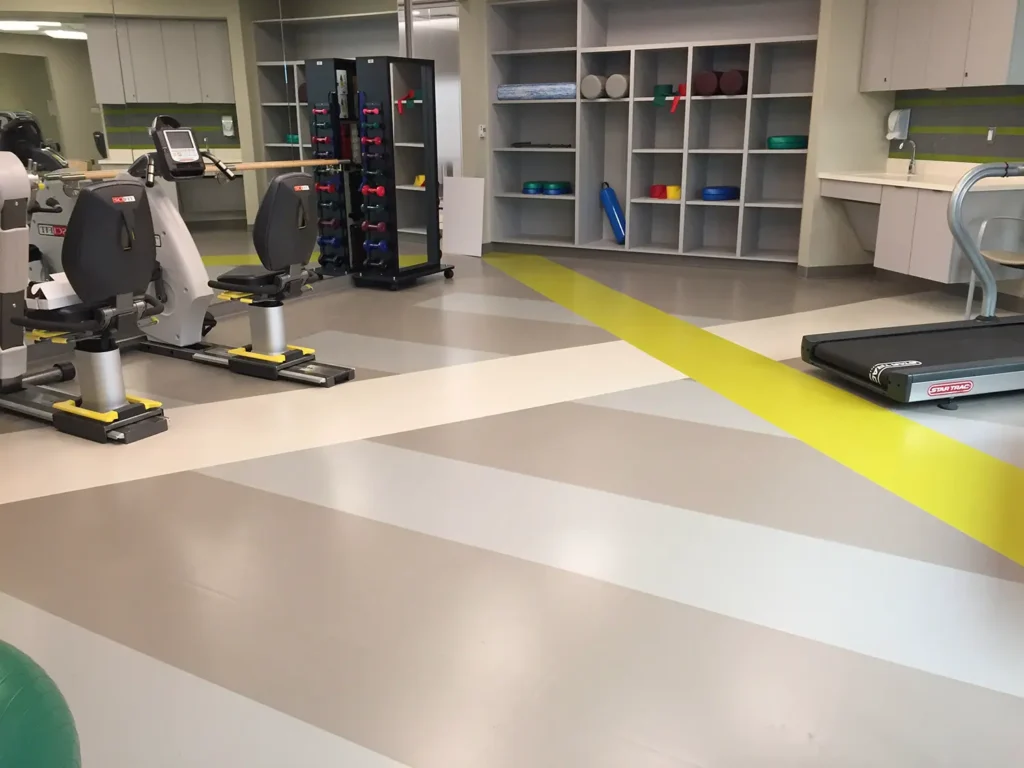 A modern physical therapy room with two exercise bikes, a treadmill, racks of dumbbells, and shelves holding mats and foam rollers, all on a striped beige and yellow floor.