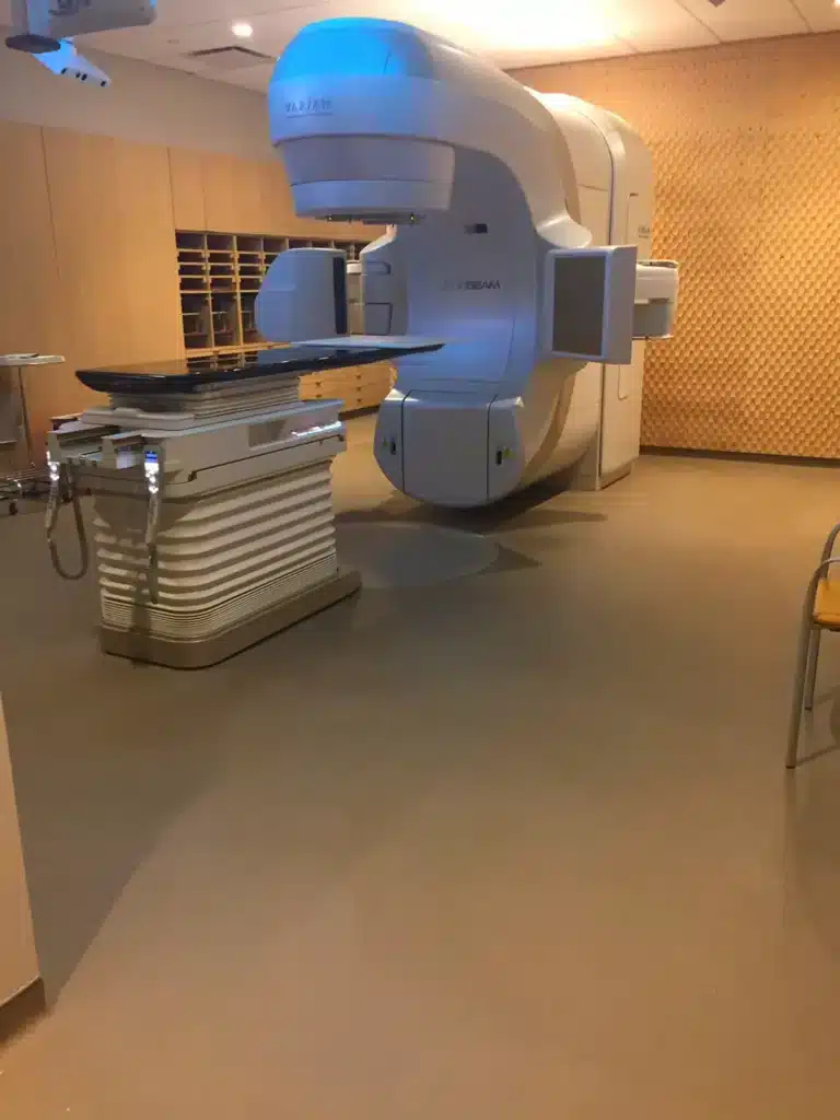 A large medical linear accelerator machine is positioned in a treatment room with beige walls and floor, used for delivering radiation therapy to patients.