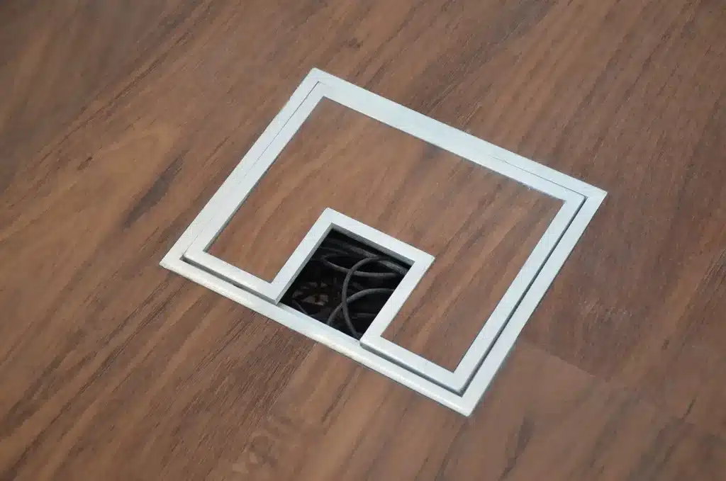A rectangular floor outlet cover with a cutout in the middle, revealing black electrical cables underneath, set in a wooden floor.