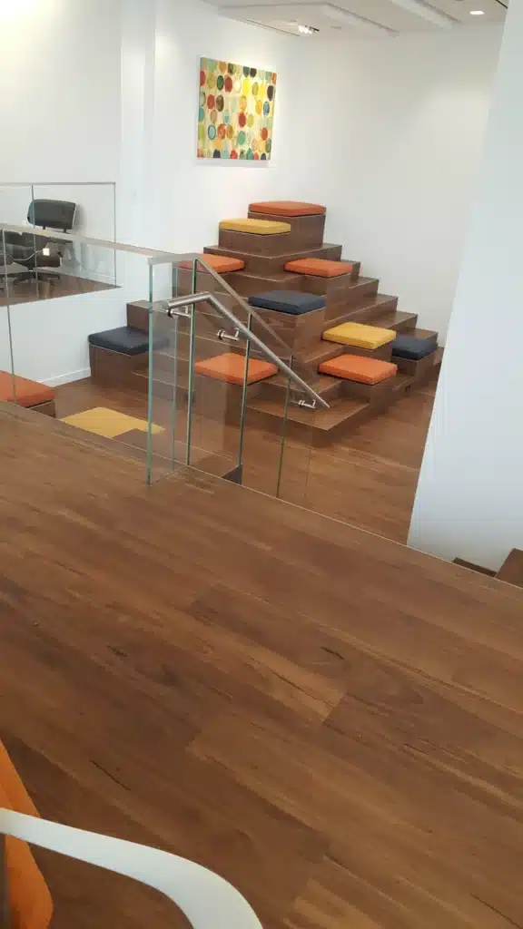 A modern indoor space with wooden floors, tiered seating with colorful cushions, glass railing, abstract art on the wall, and a chair in the background.