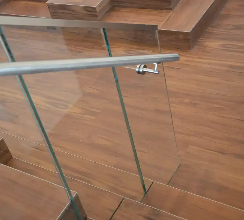 Close-up of a modern staircase with wooden steps, glass panels as the railing, and a sleek metal handrail, all set against a polished wooden floor.