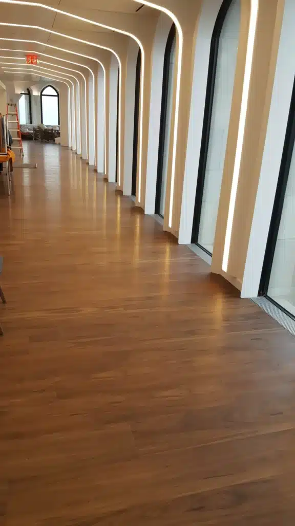 A modern hallway with wood flooring, tall arched windows, and vertical LED strip lighting along the walls. The hallway is empty and bright, with tables and chairs visible in the distance.