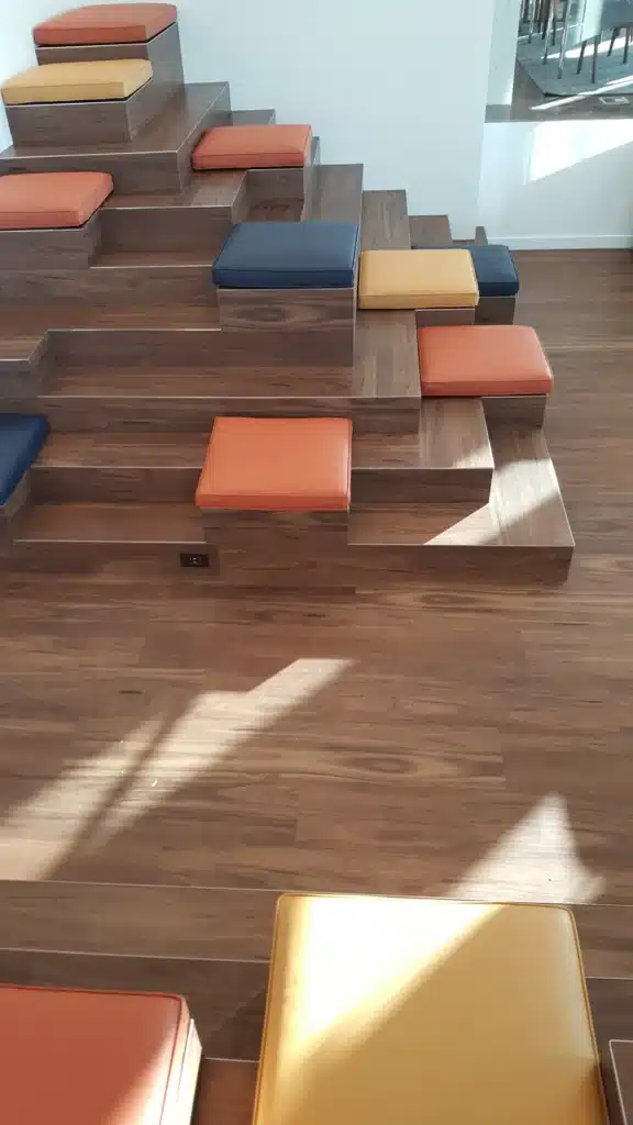 A tiered wooden seating structure with square steps, each topped with colorful cushions in orange, navy blue, and yellow. The floor and structure are made of matching wood, with sunlight casting shadows across the scene.