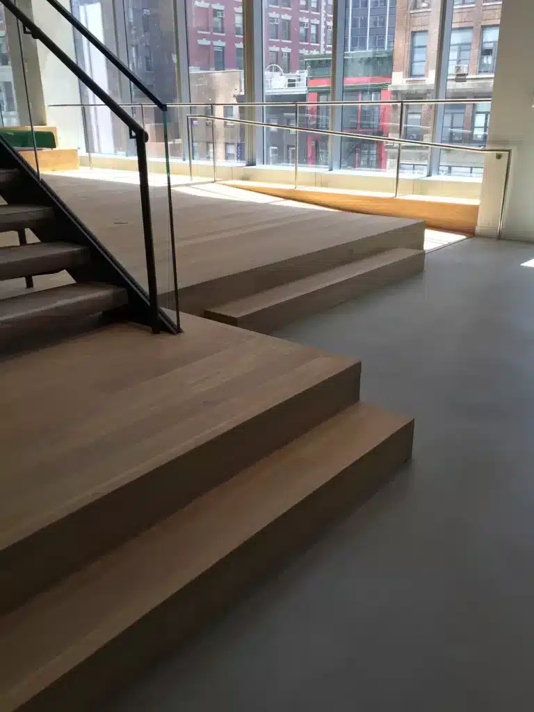 Modern interior with wide wooden stairs leading to a raised platform next to large floor-to-ceiling windows, letting in natural light and offering a view of city buildings outside.