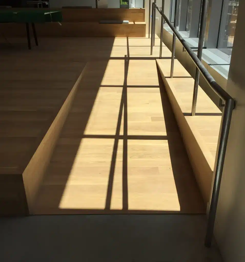Sunlight streams through tall windows, casting a grid-like shadow from a metal railing onto a wooden ramp and steps in a modern, indoor space.