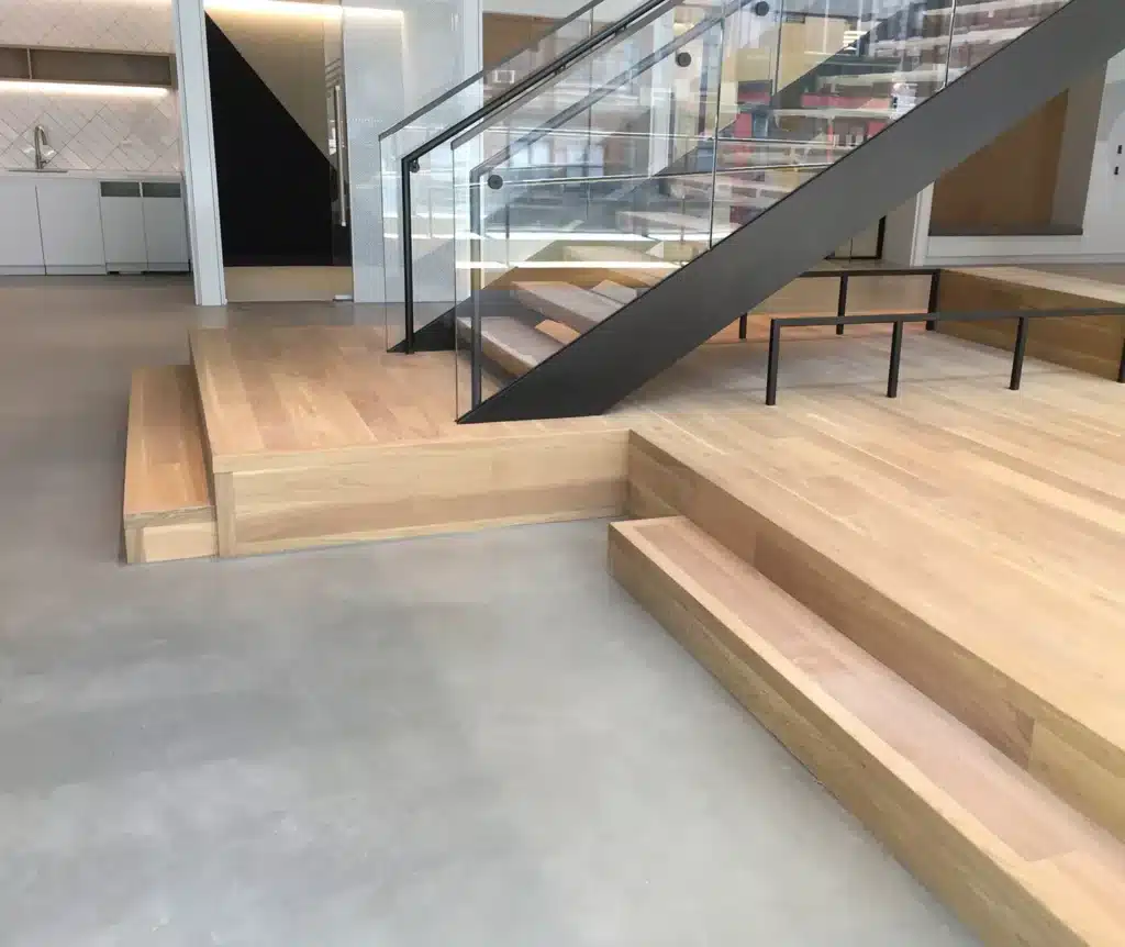 A modern interior with light wood flooring, multi-level steps, a floating staircase with glass railing, and minimalistic design elements. The area appears clean and open with neutral colors.