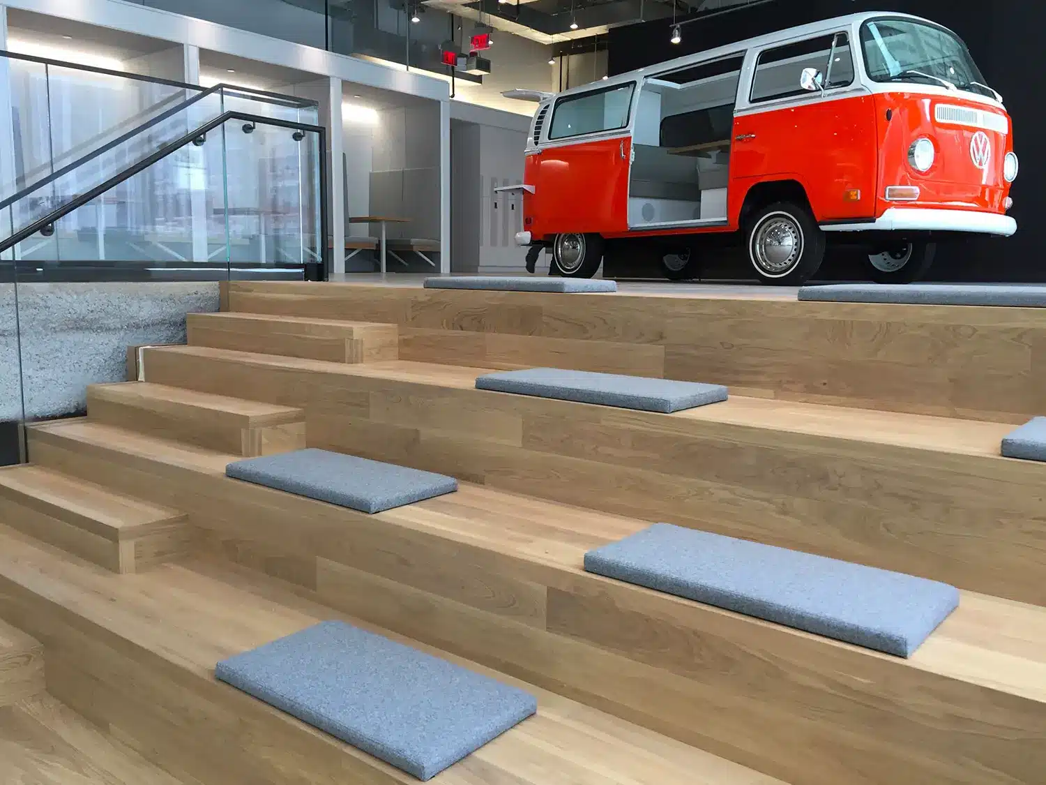 Modern office space with wooden tiered seating featuring gray cushions. An orange and white vintage van is displayed indoors near glass walls and a staircase with a glass railing.