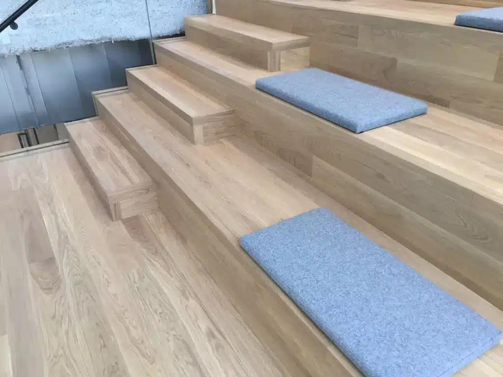 Wide wooden stairs with alternating step heights and built-in gray seat cushions; a glass railing is partially visible on the left side.
