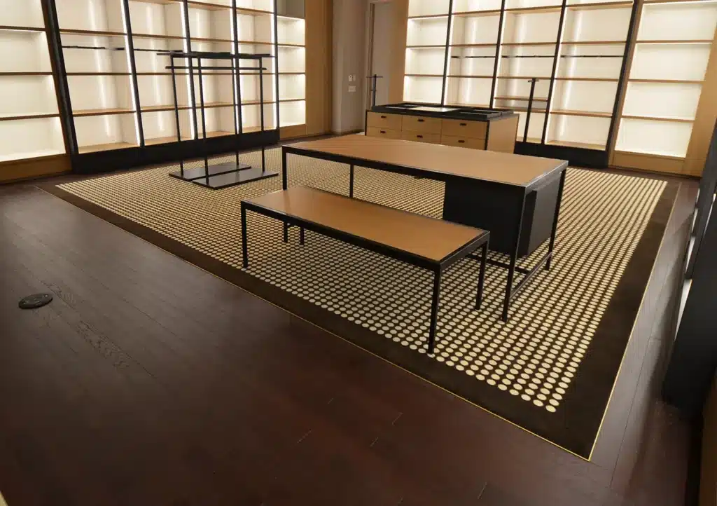 A modern, empty retail store interior with wooden floors, illuminated wall shelves, black metal racks, and a central display table and bench on a patterned, lit-up area rug.
