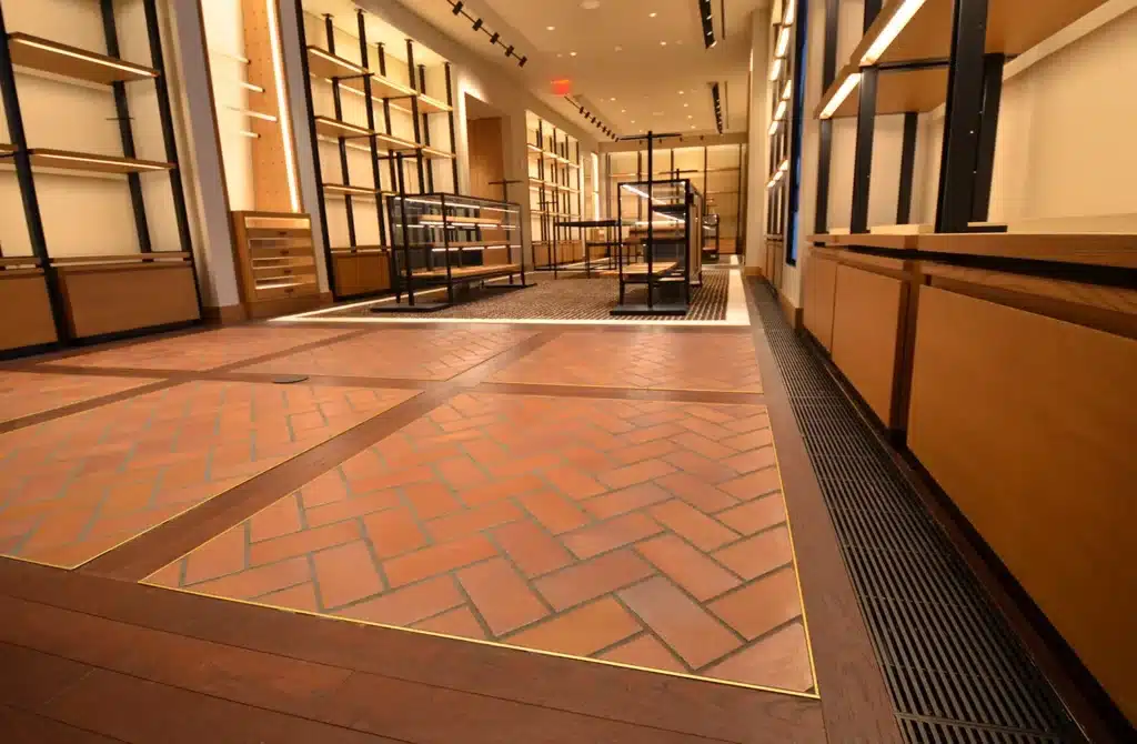 A spacious, modern retail store interior with empty wooden shelves, herringbone-patterned tile flooring, and track lighting on the ceiling. The store appears clean and well-lit, ready for merchandise.
