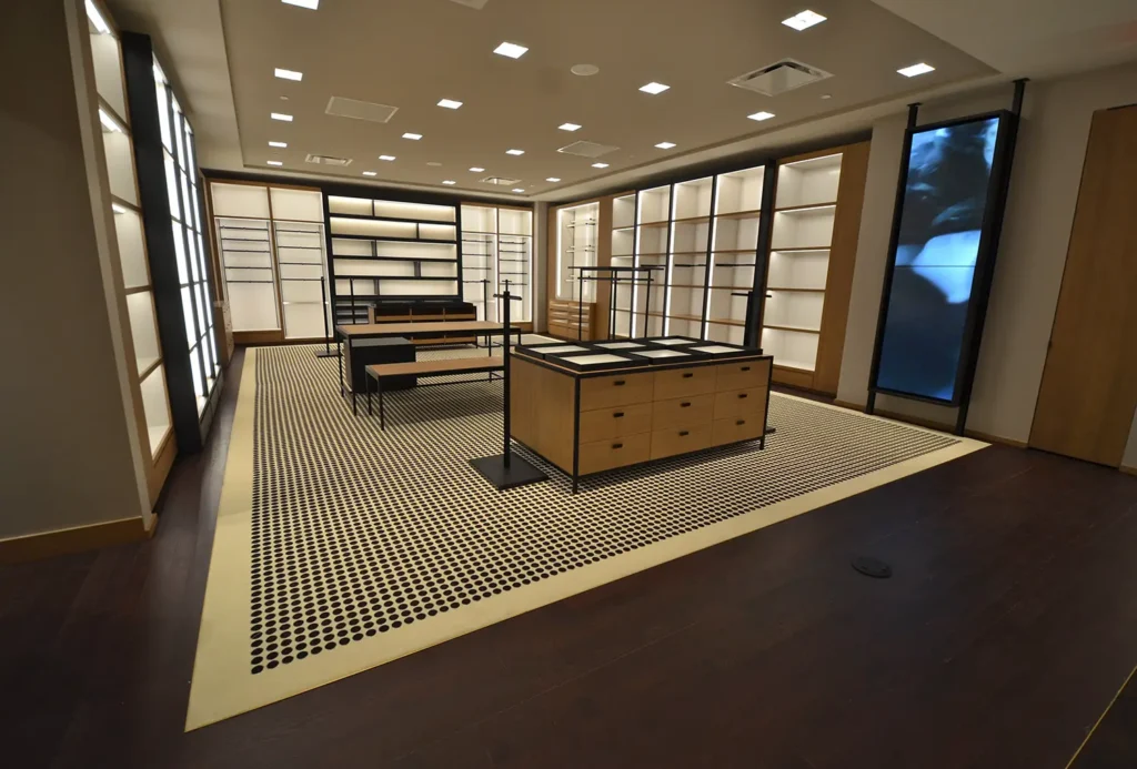 A modern, empty retail store interior with wooden display shelves, drawers, tables, and benches. The room features patterned carpet, recessed lighting, and dark wood flooring. The overall design is clean and minimalist.
