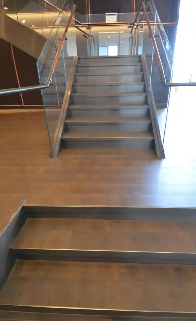 A modern indoor staircase with a small set of steps leading to a larger, wide staircase with glass railings and wooden floors, seen in a well-lit building interior.