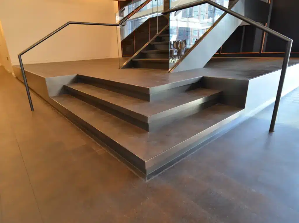 A set of three wide, shallow steps with metal edges leads up to a raised platform with modern black railings and a glass barrier, set in a contemporary indoor environment.